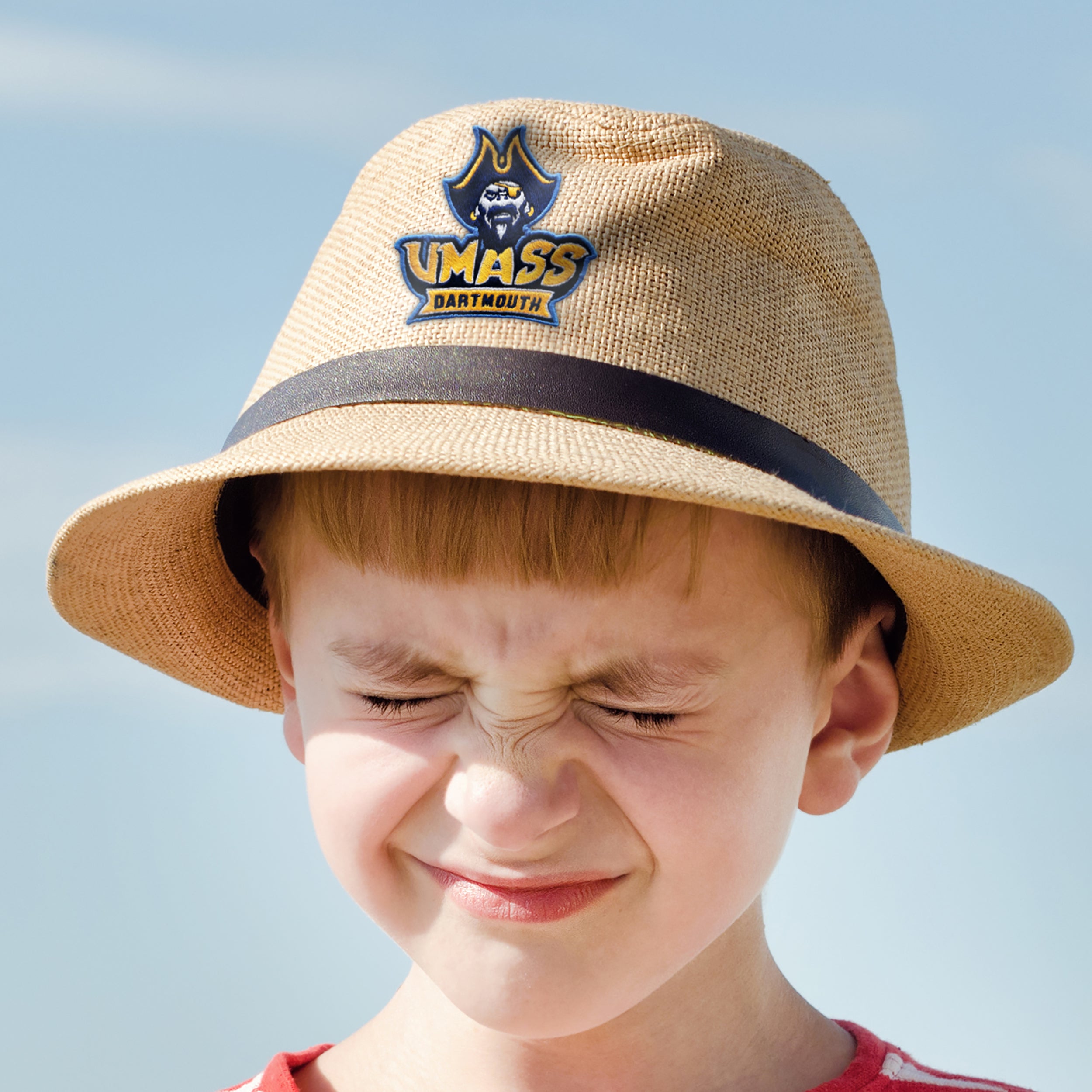 Boy wearing a straw hat with UMass Dartmouth logo, squinting in the sun outdoors.