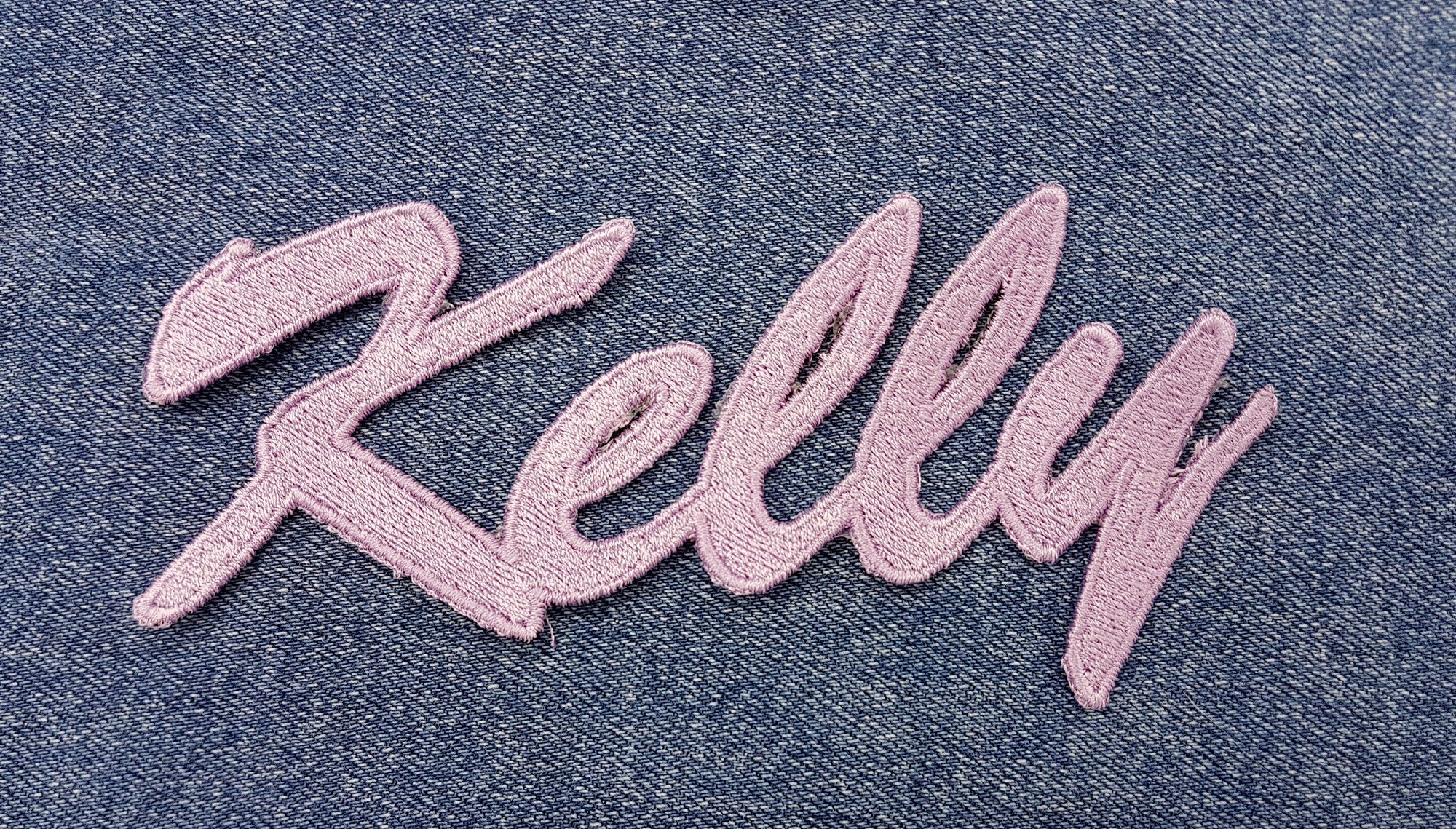 Custom embroidered name patch "Kelly" in pastel pink on denim background.