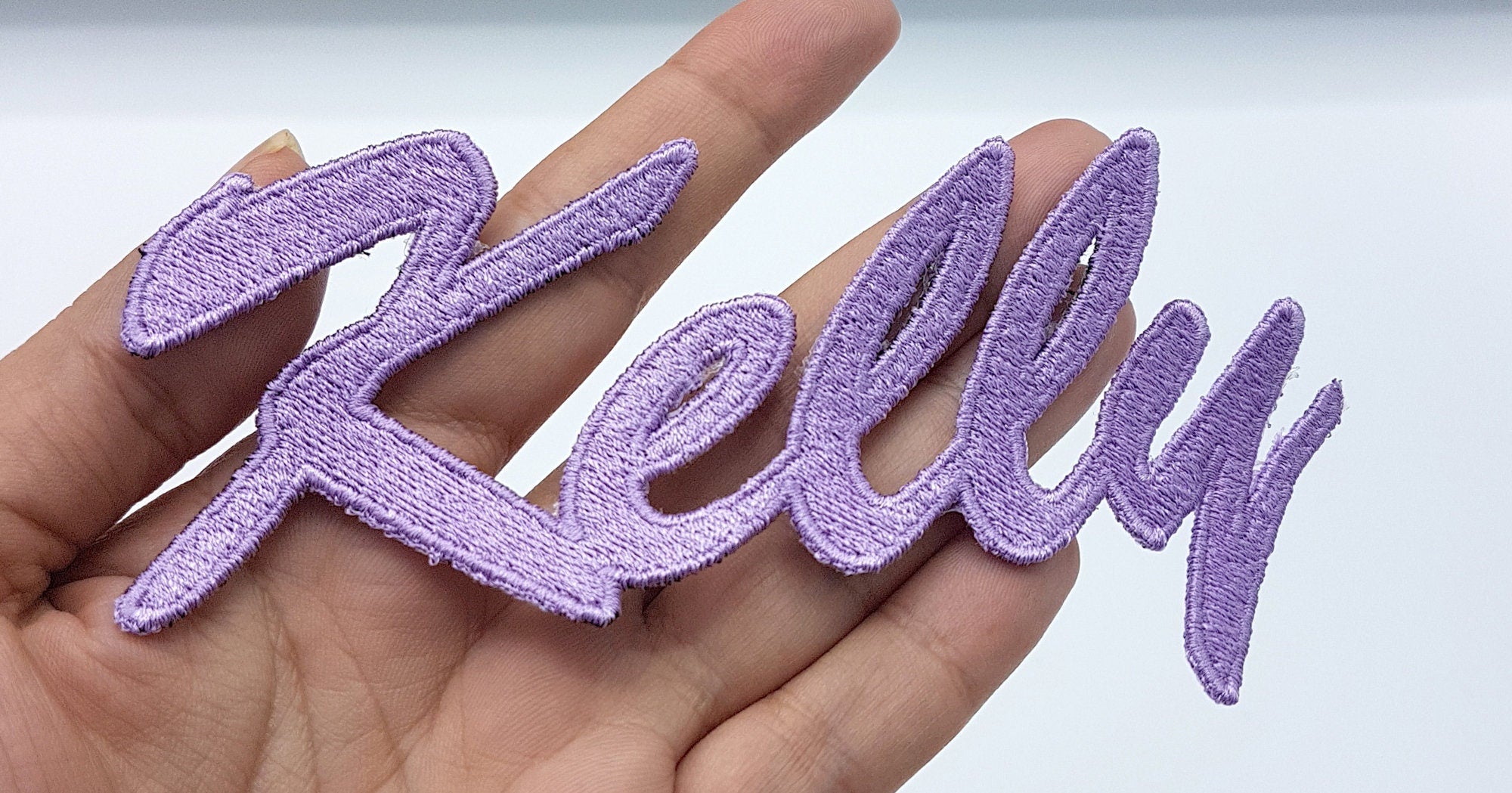 Purple embroidered name patch 'Kelly' held in a hand, custom applique for swimwear and sportswear.