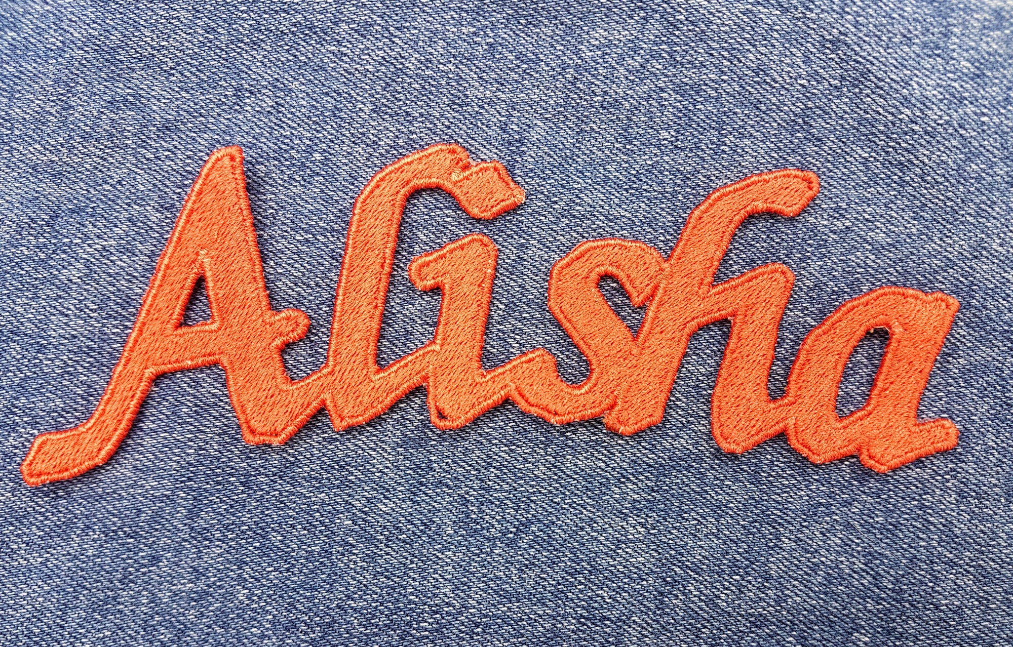 Embroidered personalized name patch reading 'Alisha' in vibrant orange on denim fabric.