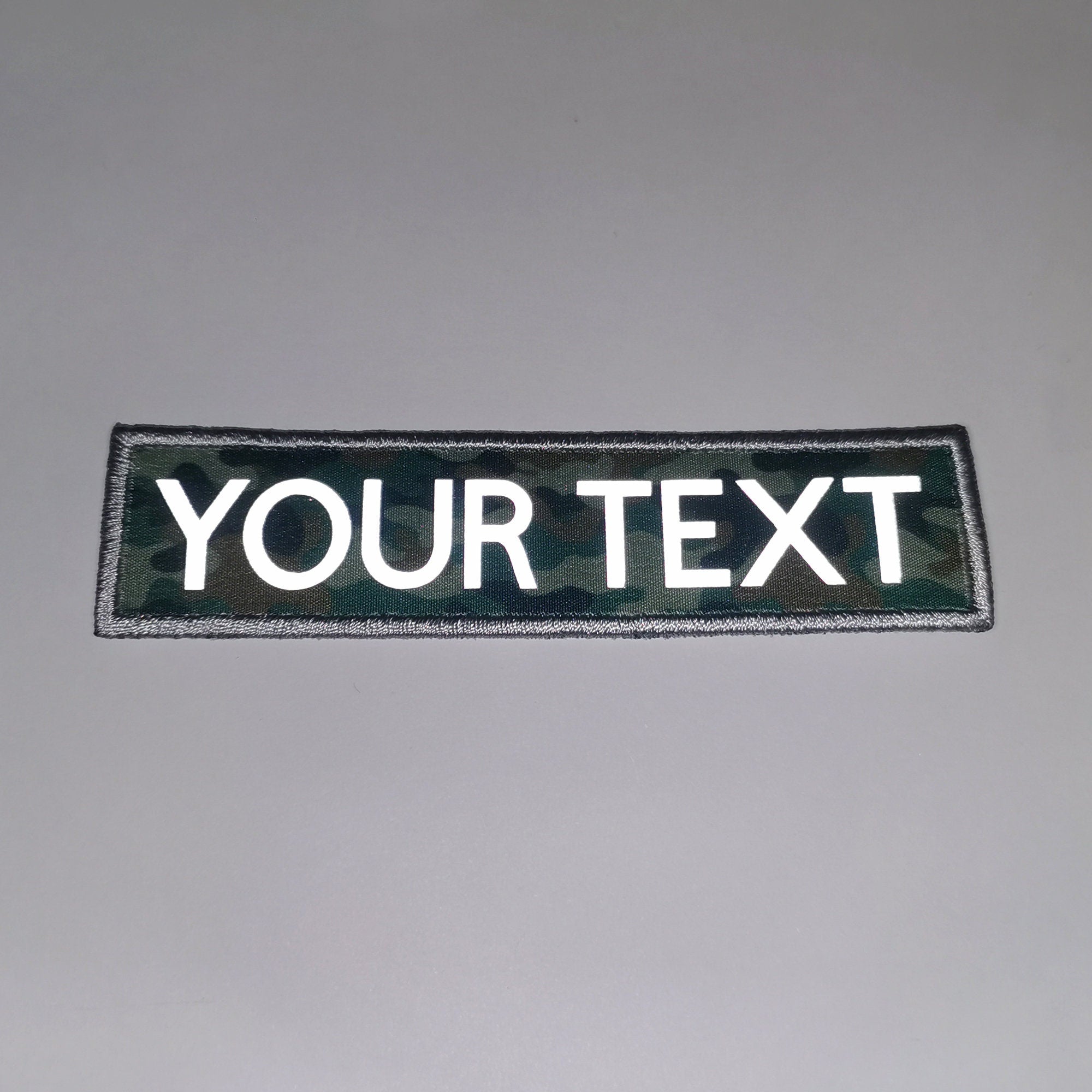 Custom reflective iron-on patch with embroidered border and printed letters on a camouflage background.
