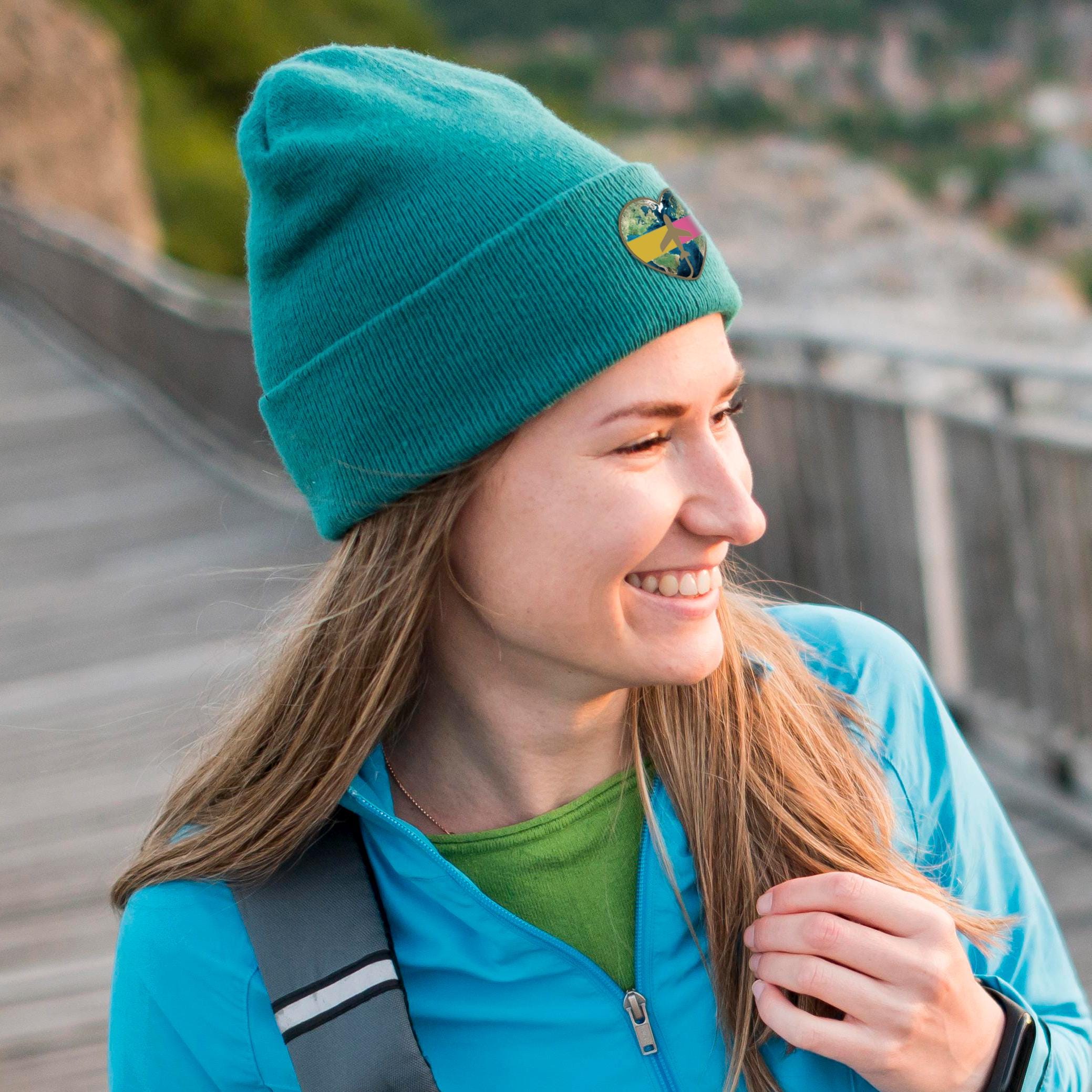 Smiling woman wearing a teal personalized beanie with a logo patch, styled with a blue jacket outdoors.