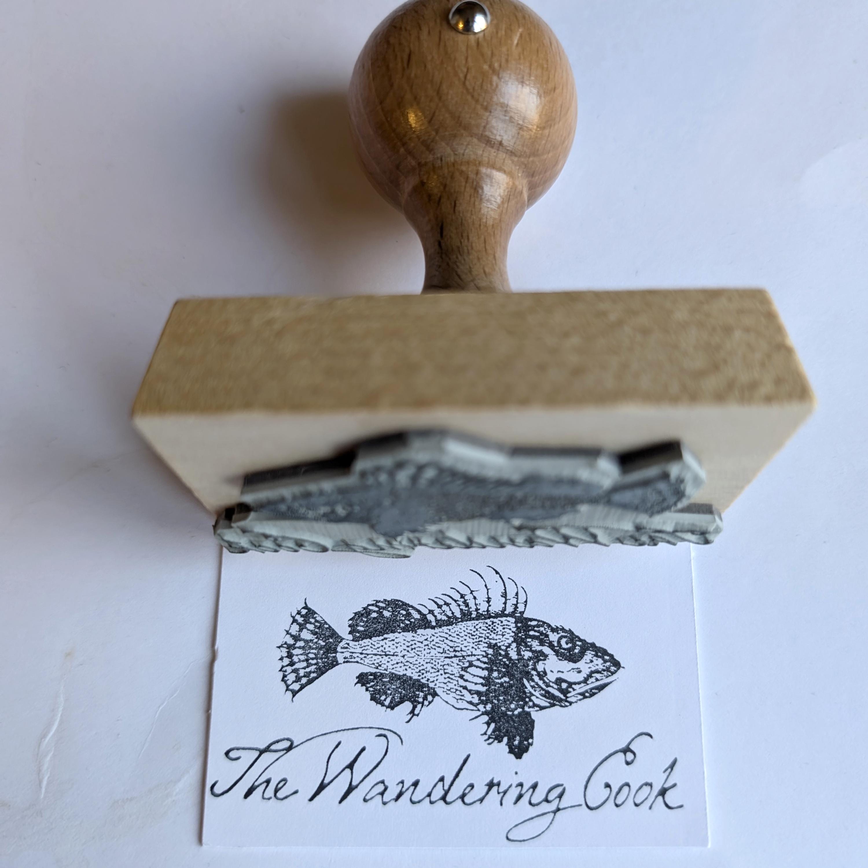 Custom logo stamp with wooden handle printing 'The Wandering Cook' on paper, ideal for branding and artwork.