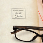Personalized library book stamp showcasing 'from the library of Clarke' next to reading glasses.