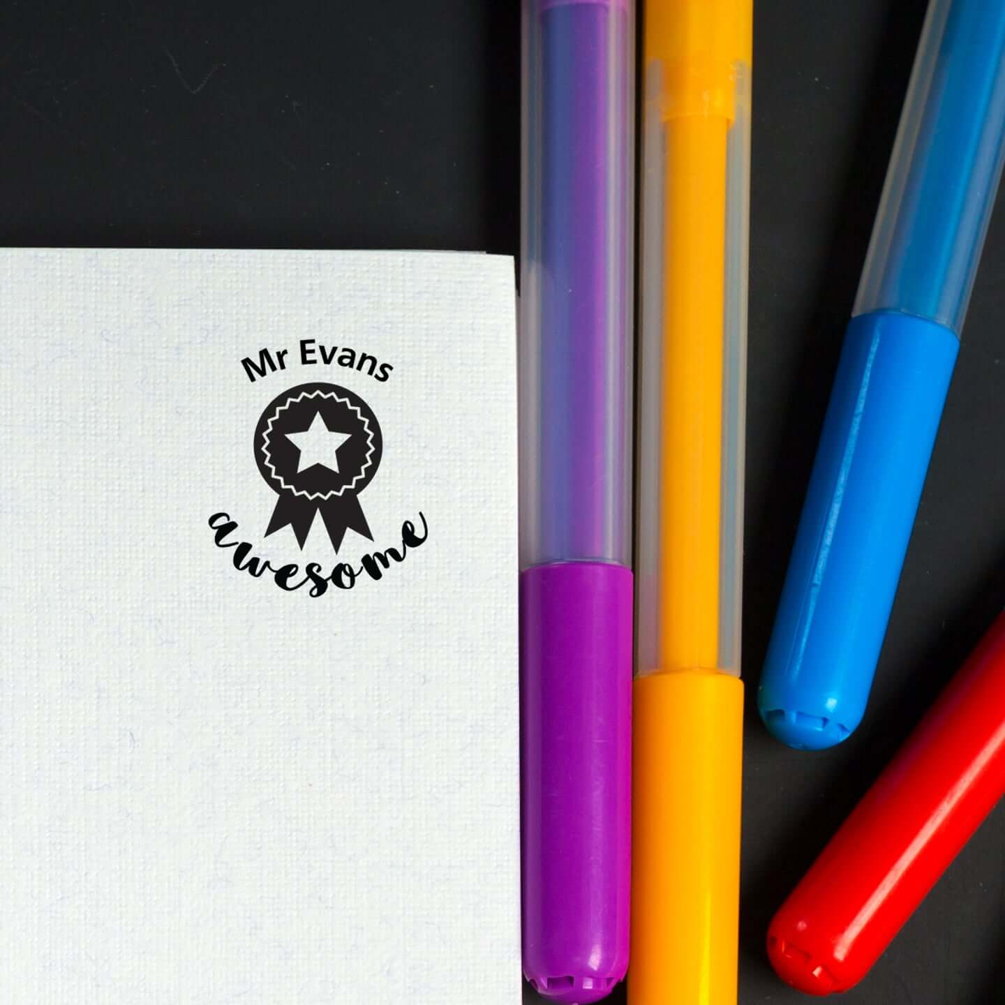 Custom teacher stamp featuring 'Mr Evans awesome' design beside colorful markers on a notebook page.