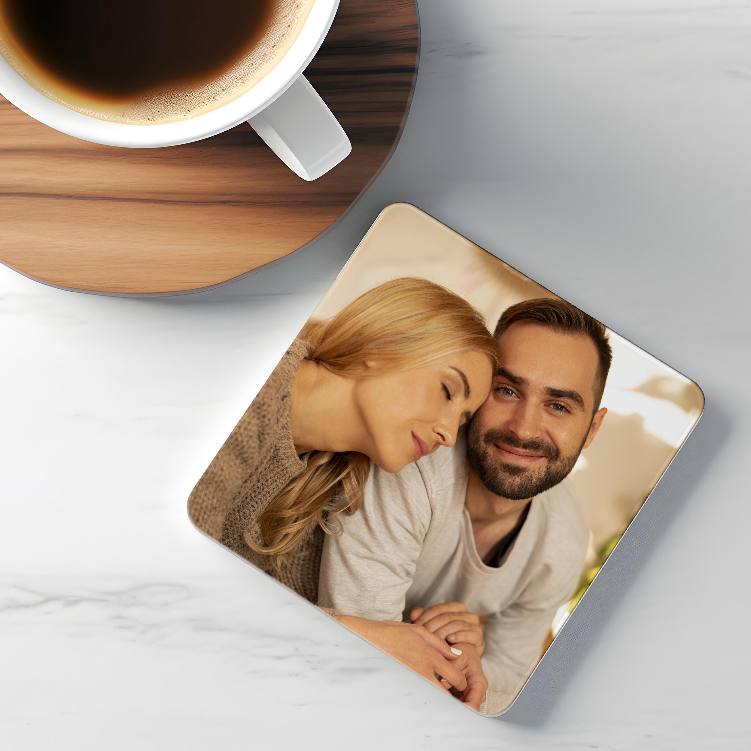 A square coaster with a photo of a couple resting their heads on each other's shoulders, accompanied by a cup of coffee on a wooden coaster.