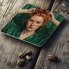 a painting of a woman with red hair, wearing a green dress, and her hands are raised to her head. coaster