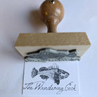 Custom logo stamp with wooden handle printing 'The Wandering Cook' on paper, ideal for branding and artwork.
